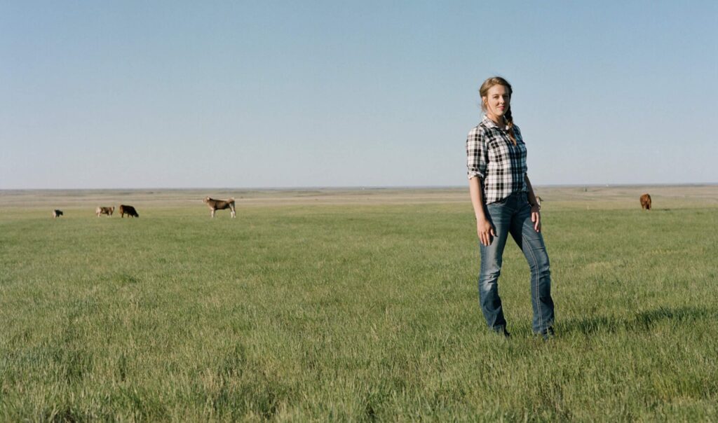 A women with blonde hair standing outside in a pasture with blue sky and cows in the distance.