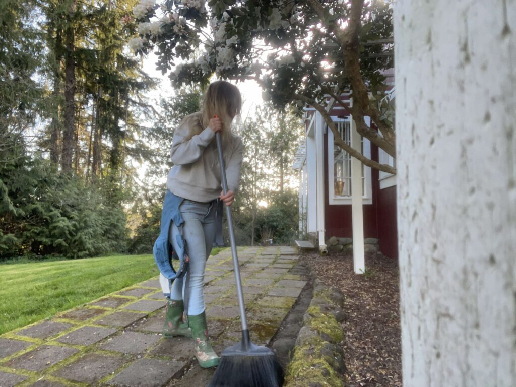 Woman sweeping sidewalk entry way on a sunny day with the sun peaking through the trees.