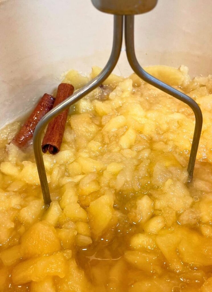 Close-up view of apples simmering on the stove after about ten minutes with cinnamon stick and potato masher to help break the apples down.