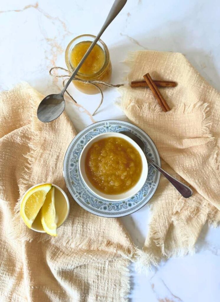 Bird's eye view of my traditional homemade cinnamon applesauce with a bowl and jar in view, plus the sliced lemon and cinnamon sticks.