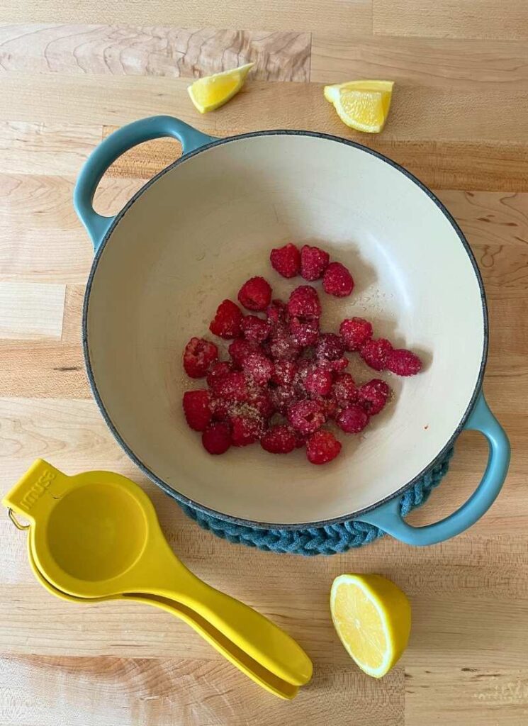 Fresh raspberries in a heavy bottomed pot with raw sugar and lemon juice squeezed on top.